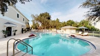 This single-story property showcases a standout outdoor amenity in the form of a large, freeform swimming pool that serves as the centerpiece of the exterior space. The pool's clear turquoise waters are bordered by a light-toned pool deck, offering ample space for lounging and recreation. Decorative planters with vibrant tropical flowers add color and visual interest along the pool's curved edges, enhancing the resort-like atmosphere.

Adjacent to the pool is a well-appointed cabana or utility structure with a hip roof, finished in a warm neutral tone that complements the surrounding architecture. A white perimeter fence encloses the pool area, providing both safety and a clean, defined boundary. The grounds are enriched by a lush canopy of mature trees that rise in the background, offering natural shade and privacy while creating a tranquil, secluded ambiance.

The main building, visible in the background, features a multi-level design with a balcony, neutral exterior finishes, and classic architectural detailing. The overall setting conveys a well-maintained, landscaped environment ideally suited for outdoor living and leisure. The combination of the expansive pool, thoughtful greenery, and structured outdoor spaces makes this property's exterior a defining and functional feature of the space.