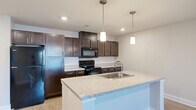 Located at 1110 Shady Brook Ln in West Haven, Utah, this single-floor, one-bedroom, one-bathroom unit offers a thoughtfully designed layout with modern finishes throughout. The open-concept kitchen and living area serves as the heart of the space, anchored by a central island with a granite-look countertop, an undermount sink, and stylish dual pendant lights overhead. Rich, dark-toned shaker-style cabinetry lines the kitchen walls, complemented by matching lower cabinets, light stone countertops, and black appliances including a range, microwave, and refrigerator. Light wood-look flooring flows through the main living area, enhancing the bright, airy feel supported by recessed lighting and a ceiling fan. The living room provides an open, versatile space with natural light from a window. The bedroom is generously sized, finished with plush carpet, a ceiling fan, and direct access to both the closet and the full bathroom. The bathroom features wood-look tile flooring, a dark vanity with a white countertop, a walk-in glass shower, and clean, neutral walls. Throughout, the unit maintains a consistent, contemporary aesthetic with soft gray tones and clean white trim, creating a cohesive and comfortable living environment.