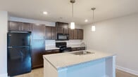 Located at 1110 Shady Brook Ln in West Haven, Utah, this single-floor, one-bedroom, one-bathroom unit offers a thoughtfully designed layout with modern finishes throughout. The open-concept kitchen and living area serves as the heart of the space, anchored by a central island with a granite-look countertop, an undermount sink, and stylish dual pendant lights overhead. Rich, dark-toned shaker-style cabinetry lines the kitchen walls, complemented by matching lower cabinets, light stone countertops, and black appliances including a range, microwave, and refrigerator. Light wood-look flooring flows through the main living area, enhancing the bright, airy feel supported by recessed lighting and a ceiling fan. The living room provides an open, versatile space with natural light from a window. The bedroom is generously sized, finished with plush carpet, a ceiling fan, and direct access to both the closet and the full bathroom. The bathroom features wood-look tile flooring, a dark vanity with a white countertop, a walk-in glass shower, and clean, neutral walls. Throughout, the unit maintains a consistent, contemporary aesthetic with soft gray tones and clean white trim, creating a cohesive and comfortable living environment.
