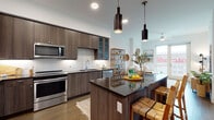 This contemporary single-floor residence showcases a thoughtfully designed open-concept layout that seamlessly connects the kitchen, dining, and living areas. The chef's kitchen is a standout feature, equipped with dark wood-grain cabinetry, a striking dark granite island with seating, a white subway tile backsplash, and a full suite of stainless steel appliances including a range, microwave, dishwasher, and French door refrigerator. Sleek cylindrical pendant lights hang above the island, adding a modern, industrial touch. Rich hardwood flooring flows throughout the main living areas, complementing the warm wood tones of the cabinetry. The living room offers a bright, airy atmosphere with clean white walls, natural light, and an outdoor view, creating an inviting space for relaxation. The bedroom is a serene retreat, featuring large windows that frame urban views and plush carpeting underfoot. The primary bathroom exudes a spa-like quality, with large-format marble-look tile flooring, a glass-enclosed walk-in shower, dark wood vanity cabinetry, and dark stone countertops. A generously sized walk-in closet with a fully customized organizational system—including shelving, hanging rods, and pull-out drawers—rounds out this well-appointed residence.