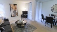 This single-floor residence offers a clean, modern aesthetic defined by bright, open interiors and thoughtful design details throughout. The living area serves as the heart of the space, featuring plush wall-to-wall carpeting that adds warmth underfoot. Large floor-to-ceiling windows dressed with white vertical blinds allow generous amounts of natural light to filter in, creating an airy and welcoming atmosphere. The living room is well-appointed, centered around a sleek circular coffee table with a glass top set atop a textured area rug, complementing the contemporary furnishings. A low-profile media console with bold hardware anchors the entertainment area, while a striking abstract artwork on the wall adds a bold visual accent. Neutral white walls throughout provide a versatile backdrop that enhances the sense of openness. The layout flows naturally, offering functional living space with a balanced blend of comfort and style. The minimalist furnishings and cohesive color palette of blacks, whites, and soft neutrals give the space a polished, sophisticated character suited to modern living.