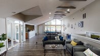 Located on Breakwater Drive in Indianapolis, Indiana, this single-story community clubhouse is designed to serve as a welcoming hub for residents and guests. The main gathering area is anchored by soaring vaulted ceilings adorned with striking exposed dark beams, recessed lighting, and rich wood-look flooring that runs throughout the space. A dramatic floor-to-ceiling window wall at one end floods the interior with natural light and offers views of the surrounding greenery, creating an open, airy atmosphere. The lounge area is furnished with comfortable gray sofas and accent chairs arranged around a glass coffee table, accented by nautical-themed décor including ship wheel motifs and coastal wall art that give the space a cohesive, relaxed character. A stately stone fireplace with a full mantle anchors one side of the room, accompanied by a cozy seating nook, a wall-mounted bulletin board, and ample open wall space. The game area features a pool table and foosball table, ideal for recreation and socializing. A separate dining nook provides an intimate setting with a round marble-top table and chairs. The pass-through kitchen includes wood cabinetry, a stainless steel refrigerator, and counter seating, offering convenient amenities for gatherings and events.