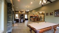 This single-floor space is designed for entertaining, offering a generous open-concept layout that seamlessly connects multiple functional zones. At the forefront is a full-sized pool table with a classic wood frame, setting the tone for the recreational nature of the space. The large-format tile flooring extends throughout, providing a cohesive and durable foundation.

The lounge area features comfortable leather seating arranged around a stone-clad fireplace, with a wall-mounted television above — creating an inviting focal point for gathering. Decorative stone columns frame the transition between zones, adding architectural character and warmth.

The kitchen and bar area is well-appointed with rich wood cabinetry, a wraparound bar with multiple bar stools, pendant lighting, and what appears to be full appliances — making it fully functional for hosting. Multiple chandeliers and recessed lighting illuminate the space, complemented by elevated ceilings that enhance the sense of openness. Natural light enters through several windows and a glass door, further brightening the interior. Overall, the space presents a refined yet casual ambiance suited for social gatherings.