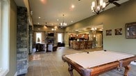 This single-floor space is designed for entertaining, offering a generous open-concept layout that seamlessly connects multiple functional zones. At the forefront is a full-sized pool table with a classic wood frame, setting the tone for the recreational nature of the space. The large-format tile flooring extends throughout, providing a cohesive and durable foundation.

The lounge area features comfortable leather seating arranged around a stone-clad fireplace, with a wall-mounted television above — creating an inviting focal point for gathering. Decorative stone columns frame the transition between zones, adding architectural character and warmth.

The kitchen and bar area is well-appointed with rich wood cabinetry, a wraparound bar with multiple bar stools, pendant lighting, and what appears to be full appliances — making it fully functional for hosting. Multiple chandeliers and recessed lighting illuminate the space, complemented by elevated ceilings that enhance the sense of openness. Natural light enters through several windows and a glass door, further brightening the interior. Overall, the space presents a refined yet casual ambiance suited for social gatherings.