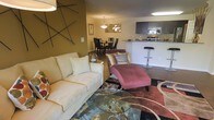 This single-floor residence offers a well-designed open-concept layout that seamlessly connects the main living areas, creating a cohesive and functional living environment. The living room serves as the centerpiece of the space, featuring a plush cream sofa, a chaise lounge in rich burgundy upholstery, and a glass-topped coffee table resting on a bold, patterned area rug. An accent wall in warm olive gold adds depth and character, adorned with an artistic branch-like wall decoration that gives the room a contemporary flair. Natural and ambient lighting work together to create a welcoming atmosphere throughout. The dining area transitions smoothly from the living room, anchored by a dark wood table and matching chairs beneath a statement pendant light fixture. Beyond the dining space, the kitchen is outfitted with dark cabinetry, light countertops, and modern appliances, complemented by a breakfast bar with sleek bar stool seating — ideal for casual dining or entertaining. The consistent use of warm tones, hardwood-style flooring, and thoughtful furnishings throughout the space creates a cohesive, inviting ambiance that balances comfort with modern style.