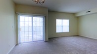 This single-floor space offers a clean, open layout designed for comfortable living. The main living area is a standout feature, bathed in natural light streaming through a large sliding glass door fitted with vertical blinds, which provides a direct view of mature trees outside, creating a serene, nature-connected atmosphere. A secondary window on the adjacent wall further enhances the brightness of the room, ensuring the space feels airy and welcoming throughout the day. The living area is finished with neutral wall-to-wall carpeting in a soft tone that complements the warm beige walls, creating a cohesive and understated aesthetic. An elegant chandelier overhead adds a decorative touch, lending a sense of refinement to the otherwise simply appointed room. The smooth ceiling and clean wall lines contribute to the open, uncluttered feel of the space. The overall ambiance is calm and neutral, making it a versatile canvas ready to accommodate a variety of interior design preferences. The connection to outdoor greenery visible through the generous glazing is a particularly notable characteristic, providing a pleasant backdrop regardless of the season.