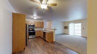 This single-floor space is designed with an open-concept layout that connects the kitchen and living area in a practical and cohesive arrangement. The kitchen is well-equipped with stainless steel appliances, including a refrigerator, electric range, microwave, and dishwasher, all complemented by warm honey-toned cabinetry and granite-look countertops. A peninsula counter serves as a natural divider between the kitchen and the adjacent living space, offering additional prep area and a casual dining option. The kitchen features durable hardwood-style flooring, while the living area transitions to plush carpeting, creating a comfortable and inviting atmosphere. A ceiling fan with an integrated light fixture is centrally positioned over the main living space, providing both airflow and ambient lighting. The living area is bright and airy, benefiting from a sliding glass door that invites natural light and provides access to an outdoor area. A wall-mounted air conditioning unit and baseboard heating ensure year-round comfort. The overall design is clean and functional, making efficient use of the available space while maintaining an open and welcoming feel throughout.