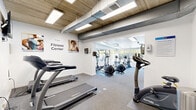 The Renew Fitness Center at 537 Tranquil Ct in Odenton, Maryland is a thoughtfully designed workout facility that combines functionality with an energizing atmosphere. The space is divided into two distinct zones seamlessly connected by an open passage, creating a cohesive and efficient layout for exercise.

The first zone features an exposed concrete and wood-panel ceiling with industrial-style ductwork and recessed panel lighting, lending the area a modern, urban aesthetic. This section houses multiple treadmills and a stationary bike, with motivational wall graphics and fitness-themed artwork adding character to the bright white walls. A wall-mounted television keeps members entertained during cardio sessions.

The second zone opens into a brighter, more expansive area bathed in natural light streaming through large floor-to-ceiling windows that offer views of an outdoor pool and lush greenery. This section is home to elliptical trainers, upright and recumbent bikes, a dumbbell rack with a full range of free weights, medicine balls, and an adjustable workout bench — providing a comprehensive strength and cross-training area.

Throughout the facility, durable rubber-backed carpet flooring provides comfort and safety underfoot. The overall ambiance is clean, welcoming, and well-lit, making it a practical and motivating environment for residents looking to maintain an active lifestyle.