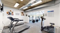 The Renew Fitness Center at 537 Tranquil Ct in Odenton, Maryland is a thoughtfully designed workout facility that combines functionality with an energizing atmosphere. The space is divided into two distinct zones seamlessly connected by an open passage, creating a cohesive and efficient layout for exercise.

The first zone features an exposed concrete and wood-panel ceiling with industrial-style ductwork and recessed panel lighting, lending the area a modern, urban aesthetic. This section houses multiple treadmills and a stationary bike, with motivational wall graphics and fitness-themed artwork adding character to the bright white walls. A wall-mounted television keeps members entertained during cardio sessions.

The second zone opens into a brighter, more expansive area bathed in natural light streaming through large floor-to-ceiling windows that offer views of an outdoor pool and lush greenery. This section is home to elliptical trainers, upright and recumbent bikes, a dumbbell rack with a full range of free weights, medicine balls, and an adjustable workout bench — providing a comprehensive strength and cross-training area.

Throughout the facility, durable rubber-backed carpet flooring provides comfort and safety underfoot. The overall ambiance is clean, welcoming, and well-lit, making it a practical and motivating environment for residents looking to maintain an active lifestyle.