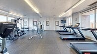 Located at 1100 Penn Center Blvd in Pittsburgh, PA, this well-appointed fitness center offers a comprehensive workout environment across a single open floor. The space is thoughtfully laid out to accommodate a wide range of exercise needs, from cardiovascular training to strength conditioning. Durable rubber tile flooring runs throughout, providing both safety and comfort underfoot.

The cardio section is equipped with multiple treadmills, elliptical trainers, and stationary bikes, including True Fitness branded equipment. Several flat-screen televisions are mounted throughout the space, allowing users to stay entertained during their workouts. The strength training area features a multi-function cable machine, a weight bench, and a dumbbell rack stocked with a variety of free weights. Stability balls and additional accessories are also available for functional training.

Large windows line one side of the fitness center, flooding the space with natural light and offering sweeping views of the Pittsburgh skyline and surrounding landscape — a motivating backdrop for any workout. Recessed linear lighting along the ceiling supplements the natural light, ensuring the space remains bright and functional at all hours. Framed motivational artwork decorates the white walls, contributing to an energizing yet clean aesthetic. The overall ambiance is professional, well-maintained, and welcoming to users of all fitness levels.