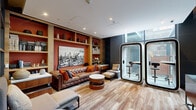 This sophisticated lounge space masterfully balances warmth and contemporary design across a single open floor. The room is anchored by a rich, tufted brown leather Chesterfield sofa accented with vibrant orange and blue throw pillows, set against a striking built-in shelving unit finished in warm wood tones with a terracotta-orange backdrop. The shelves display an artful arrangement of books, vases, and decorative objects, while a large black-and-white cityscape artwork serves as a bold focal point. Reclaimed-style hardwood flooring adds texture and character throughout the space, complementing the warm color palette. On the opposite end, a floor-to-ceiling glass curtain wall floods the area with natural light and offers expansive urban views of the surrounding cityscape. Two sleek, freestanding private meeting pods provide a modern, functional contrast to the lounge's residential warmth, ideal for focused work or private conversations. A rich walnut credenza with decorative lamps adds refinement near the glazed perimeter. Glass partition doors connect this space to an adjacent area, hinting at a larger, cohesive environment. The overall ambiance is one of curated comfort — a thoughtfully designed space where social relaxation and productivity coexist seamlessly.