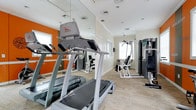 This fitness center is thoughtfully designed to provide a functional and motivating environment for exercise. The space is defined by its clean, modern aesthetic, with light wood-style flooring underfoot and a crisp white ceiling fitted with recessed lighting. A bold orange accent wall adds a vibrant, energetic touch that sets the tone for an active atmosphere.

Floor-to-ceiling mirrors line one wall, visually expanding the space and allowing users to monitor their form during workouts. The room is outfitted with a selection of cardio equipment, including treadmills and a Freemotion elliptical trainer, positioned to make the most of the available floor space. Strength training is also well represented, with a multi-station cable machine and additional resistance equipment positioned toward the rear of the room.

Natural light filters in through multiple windows, brightening the space and contributing to an open, airy feel. The layout is practical and uncluttered, ensuring ease of movement between equipment. Whether focused on cardiovascular fitness or strength building, this facility offers a well-rounded range of equipment to accommodate a variety of fitness routines in a clean, purpose-built setting.