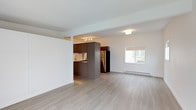 This single-floor unit offers an open-concept layout with a seamless flow between the living area and kitchen. The flooring throughout is light-toned hardwood, contributing to a clean, contemporary aesthetic that enhances the sense of space.

The living area is generously proportioned, with crisp white walls and a textured ceiling. A window along one wall allows natural light to filter into the space, brightening the interior. A wall-mounted panel, likely for climate or lighting control, is visible near the window.

The kitchen is positioned toward the rear of the open-plan space and features two-tone cabinetry with warm wood-grain lower cabinets and darker upper cabinets. A peninsula or island provides additional workspace and visual separation from the living area. The kitchen also includes overhead cabinetry and appears to be equipped with standard appliances.

A central partition wall with a built-in closet divides the main living space from a hallway, which leads to additional rooms. Frosted glass elements are visible along the corridor, allowing light to pass through while maintaining privacy. The unit's overall ambiance is bright, clean, and modern.