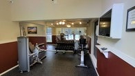 This fitness center offers a comprehensive workout environment designed to accommodate a wide range of exercise routines. The space features soaring ceilings that lend an open, airy feel, complemented by large windows that invite abundant natural light and provide views of the surrounding greenery outdoors. The walls are finished in a two-tone scheme with deep burgundy lower panels and light upper walls, creating a bold yet focused atmosphere conducive to exercise. Full-length mirrors line one wall, visually expanding the space while allowing users to monitor their form during workouts. The rubber flooring throughout provides durability and cushioning underfoot. Equipment is thoughtfully arranged to maximize usable floor space. A fully stocked dumbbell rack holds an extensive range of free weights, while multiple cardio machines — including a stationary bike and additional aerobic equipment — are positioned for easy access. Strength and resistance training machines round out the offerings. Overhead, a ceiling fan and track lighting ensure the space remains comfortable and well-lit during any workout. A counter along one wall provides a convenient surface for personal belongings. Overall, the fitness center delivers a functional, well-appointed environment suited for both cardio and strength training.