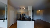 This single-floor space showcases a thoughtfully designed open-concept layout that seamlessly connects the kitchen and living areas, creating a sense of spaciousness and flow. The kitchen serves as a central hub, anchored by a generously sized island with a striking dark countertop that provides ample workspace and casual seating. Light wood cabinetry lines the walls, offering abundant storage, while stainless steel appliances — including a refrigerator, gas range, and built-in microwave — complete the modern culinary setup. A sleek tile backsplash adds a refined detail behind the range and sink. Track lighting mounted on the ceiling provides focused illumination throughout the kitchen, complementing the overall contemporary aesthetic. The flooring throughout the space features warm hardwood-style planks that add texture and continuity between rooms. The living area, visible beyond the kitchen, is bright and open, benefiting from the connected layout. Neutral wall tones keep the atmosphere clean and versatile, making the space easy to personalize. Overall, the unit presents a well-appointed, functional design with modern finishes suited to everyday living.