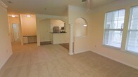 This single-floor space showcases an open-concept design that seamlessly connects the living, dining, and kitchen areas. The layout is defined by distinctive architectural details, including arched passageways with decorative columns that serve as elegant dividers between spaces, adding both structure and visual interest without enclosing the floor plan.

The living area features neutral-toned wall-to-wall carpeting, contributing to a warm and comfortable atmosphere. A ceiling fan is visible overhead, providing both functional airflow and a subtle design element. Natural light enters through a window on the right side of the living space, enhancing the bright, airy feel of the interior.

The kitchen is accessible via one of the arched openings and is equipped with stainless steel appliances, including a refrigerator and microwave. A dark-topped breakfast bar or counter separates the kitchen from the adjoining areas, creating a practical transition zone. The kitchen and adjacent areas feature hard flooring, contrasting with the carpeted living space.

A pendant light fixture is mounted within one of the arched niches, adding a decorative accent. The overall color palette remains consistent with soft, neutral tones throughout, creating a cohesive and inviting environment.