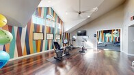 This single-floor fitness and community space combines energetic design with practical amenities. The gym area immediately draws attention with its striking bold geometric wall mural in teal, gold, rust, and navy tones, creating a dynamic and motivating environment. Soaring vaulted ceilings with a ceiling fan and arched windows flood the space with natural light, while rich hardwood flooring runs throughout. The cardio zone is equipped with stationary bikes and a recumbent bike, complemented by a wall-mounted TV for entertainment during workouts. A dedicated strength and functional training area features free weights, dumbbells, adjustable benches, stability balls, resistance equipment, and exercise mats, all arranged against another vivid geometric accent wall. Large windows provide views of the surrounding outdoors. A full-wall mirror enhances the sense of space in the weight room, which transitions to carpet flooring. Adjacent to the gym is a comfortable lounge and kitchenette area, featuring dark cabinetry with granite countertops, a stainless steel refrigerator, a sink, and a coffee station. Dining tables with modern white metal chairs, recessed lighting, a wall-mounted TV, and a window with outdoor views complete this inviting communal retreat.