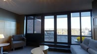 This single-floor space makes a striking first impression with its expansive floor-to-ceiling windows framed in dark metal, flooding the interior with natural light and providing unobstructed views of the surrounding urban skyline. The large glazed panels seamlessly connect the interior living area to a spacious private terrace, accessible via full-height glass doors. The terrace features a railed perimeter, open sky exposure, and outdoor seating — making it an ideal setting for relaxing or entertaining while taking in elevated city views.

Inside, the living area is thoughtfully appointed with a comfortable seating arrangement, including a sofa and armchair, centered around a contemporary low-profile coffee table. The dark carpeted flooring, deep window sills, and rich brick exterior facade visible through the glass contribute to a moody, sophisticated aesthetic. The interplay of dark tones with abundant natural light gives the space a refined, metropolitan character.

The overall design balances industrial elements — exposed brick, dark metal framing — with clean, modern furnishings, creating an environment that is both functional and visually compelling. The direct connection to the outdoor terrace further enhances the sense of openness and scale throughout.