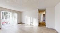 This single-floor unit presents a clean, functional layout with a neutral color palette throughout. The main living area features light wood-style flooring that extends through the common spaces, complemented by white walls and a textured ceiling. A sliding glass door fitted with vertical blinds allows abundant natural light to fill the living room while providing access to an outdoor space with views of surrounding greenery.

The kitchen is well-appointed with warm-toned wood cabinetry extending to the ceiling for ample storage, light gray laminate countertops, a stainless steel sink, a full-size refrigerator, and a dishwasher. Light wood flooring carries through this space, and a window supplies natural light. An adjacent area provides additional flexible living or dining space.

The bedrooms feature plush neutral carpeting, vertical-blind-covered windows that bring in natural light, and wall-mounted heating vents. The bathroom includes a wood-toned vanity with a countertop sink, a large mirror, and a full bathtub-shower combination, all finished with matching light wood flooring. A hallway connects the private areas, completing this straightforward and well-organized floor plan.