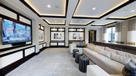 This elegantly designed single-floor space exudes a modern, upscale aesthetic anchored by its most commanding architectural feature: a deeply detailed coffered ceiling adorned with dark trim accents and integrated ambient lighting. The warm glow of recessed cove lighting within each coffer adds depth and a welcoming atmosphere, complementing the crisp white ceiling panels above.

The walls are thoughtfully designed with a structured panel system of dark-framed display sections, each showcasing framed artwork that contributes to the space's refined, gallery-like character. Recessed spotlights within these wall panels highlight the art, drawing the eye across the room.

The main lounge area is furnished with generous, cream-toned sectional and accent sofas, arranged to encourage conversation and relaxation. A set of dark, sculptural side tables with a fluted cylindrical design provide striking contrast against the light-toned carpeting underfoot, which features a subtle textured pattern that adds warmth to the overall palette.

A large flat-screen display is mounted on the left wall, enhancing the space's entertainment functionality. Natural light filters in through sheer curtains visible toward the far end, softening the interior ambiance. The overall design balances boldness and sophistication through its consistent use of a black, white, and cream color palette.