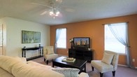 This single-floor residence offers a thoughtfully designed living space that balances comfort and style. The living room serves as the heart of the home, immediately drawing attention with its bold terracotta-orange accent wall that adds warmth and character to the room. A textured ceiling with a ceiling fan overhead provides both visual interest and practical air circulation throughout the space.

The living area is generously proportioned, comfortably accommodating a plush sofa, a pair of patterned accent chairs, and a dark wood coffee table, creating an ideal arrangement for relaxing or entertaining. A matching dark wood media console anchors the accent wall, providing ample storage and display space. Natural light filters in through multiple windows dressed with sheer curtains, lending the room a bright and airy feel.

Soft, neutral-toned carpeting runs underfoot, complementing the warm color palette of the walls and furnishings. Decorative touches, including framed wall art and fresh floral accents, contribute to the overall inviting ambiance. The cohesive design of the space reflects a careful balance of earthy tones and neutral furnishings, creating an atmosphere that feels both lively and relaxed — a welcoming environment suited for day-to-day living.