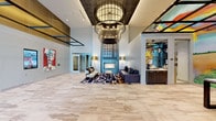 Studio 3807's lobby makes an immediate impression through its dramatic double-height ceilings and expansive, light-toned stone tile flooring that stretches throughout the space. The entrance features floor-to-ceiling glass doors framed in warm bronze tones, flanked by dark stone columns and elevator banks, establishing a polished yet edgy aesthetic from the moment of arrival. A sculptural bronze figure greets visitors near the entryway, setting the tone for the art-centric identity of the building.

The central lounge area anchors the main lobby, where a bold, oversized metal fireplace surround finished in steel blue commands attention. Flanking floor lamps in brushed gold complement a leather sofa and an abstract, graphic area rug, creating an inviting yet design-forward seating vignette. Overhead, an elaborate multi-tiered chandelier with cage-like detailing serves as a striking focal point.

Vibrant, large-scale murals and original paintings are positioned throughout, including a colorful portrait mural that dominates one dining alcove, where pendant lights hang above a communal table beneath a gridded ceiling detail. Digital information displays and additional curated artwork line the walls, reinforcing the space's gallery-like character. The overall ambiance is contemporary, bold, and culturally expressive.