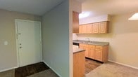 This single-floor residential space offers a practical and well-organized layout designed for comfortable everyday living. The kitchen is a standout feature, outfitted with warm light wood cabinetry that provides ample upper and lower storage. A double-basin sink sits beneath a skylight-style overhead fixture that brightens the workspace, while a dishwasher is neatly integrated beneath the counter. The countertops offer generous prep space, and a half-wall peninsula partially separates the kitchen from the adjoining area, creating an open yet defined sense of flow between spaces. The flooring transitions from dark hardwood-style planks in the kitchen to carpet in the adjacent room, adding both texture and warmth throughout. Neutral-toned walls contribute to a clean, understated aesthetic that allows for easy personalization. A hallway leading to a white-paneled door suggests additional rooms or storage beyond, reinforcing the functional nature of the floor plan. The textured ceiling and consistent lighting throughout lend the space a cohesive, move-in-ready character suited to a variety of lifestyles.