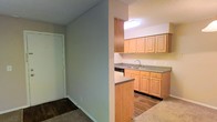 This single-floor residential space offers a practical and well-organized layout designed for comfortable everyday living. The kitchen is a standout feature, outfitted with warm light wood cabinetry that provides ample upper and lower storage. A double-basin sink sits beneath a skylight-style overhead fixture that brightens the workspace, while a dishwasher is neatly integrated beneath the counter. The countertops offer generous prep space, and a half-wall peninsula partially separates the kitchen from the adjoining area, creating an open yet defined sense of flow between spaces. The flooring transitions from dark hardwood-style planks in the kitchen to carpet in the adjacent room, adding both texture and warmth throughout. Neutral-toned walls contribute to a clean, understated aesthetic that allows for easy personalization. A hallway leading to a white-paneled door suggests additional rooms or storage beyond, reinforcing the functional nature of the floor plan. The textured ceiling and consistent lighting throughout lend the space a cohesive, move-in-ready character suited to a variety of lifestyles.