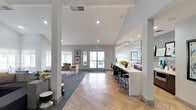This single-floor communal space showcases a thoughtfully designed open-plan layout that seamlessly blends multiple functional zones within one expansive area. Soaring ceilings with exposed white beams amplify the sense of openness, while an abundance of natural light floods the interior through large windows and glass entry doors that frame views of the surrounding outdoor areas.

The flooring transitions between herringbone-patterned hardwood and carpeted sections with teal accent strips, subtly defining separate zones within the open space. A bold teal feature wall anchors the main lounge area, which is furnished with a large sectional sofa, accent chairs, and a built-in media wall complete with mounted televisions.

A sleek counter-height bar area with black stools and pendant lighting offers an alternative workspace or social gathering spot. Adjacent to this is a well-appointed kitchenette featuring white quartz countertops, gray subway tile backsplash, a floating display shelf with curated artwork, and dark cabinetry with stainless steel fixtures.

A cozy window nook with built-in bench seating, decorative cushions, and an adjoining table provides an intimate retreat. Framed artwork throughout and carefully chosen greenery complete the polished, contemporary ambiance of this versatile communal space.