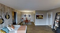 This single-floor space showcases an open-concept design that seamlessly connects the living and dining areas, creating a cohesive and functional layout. The flooring throughout is a rich, dark wood-look material that anchors the space with warmth and continuity.

The living area, positioned in the foreground, features a light-toned sectional sofa, offering a comfortable gathering spot that flows naturally into the dining space. The dining area is defined by a striking reclaimed wood accent wall in warm, natural tones, which serves as a focal point and adds rustic character. A round glass-top dining table paired with orange metal chairs sits atop a geometric black-and-white area rug, adding a contemporary contrast to the organic textures nearby. A classic pendant chandelier overhead provides ambient lighting.

Adjacent to the dining area, a pass-through window with a stone tile backsplash offers a glimpse into the kitchen, hinting at additional functionality. The neutral gray walls throughout keep the space feeling bright and versatile. Textured white ceilings and clean white baseboards complete the polished, well-maintained aesthetic of this inviting single-story space.