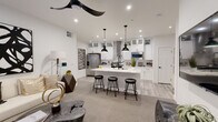 This well-appointed single-floor residence showcases a cohesive modern aesthetic defined by crisp white walls, black hardware accents, and a carefully curated monochromatic palette throughout. The open-concept living and kitchen area forms the heart of the home, where a spacious island with striking quartz countertops and black pendant lighting anchors the space. The kitchen is outfitted with floor-to-ceiling white shaker cabinetry featuring glass uppers, a stainless steel appliance suite including a French door refrigerator and built-in microwave, a tile backsplash, and a sleek range hood. Light wood-look flooring flows through the kitchen, transitioning to plush carpet in the living areas. The living room offers a comfortable and stylish retreat, highlighted by a large-scale abstract artwork, a modern sectional sofa, and a sculptural live-edge coffee table, with a black ceiling fan overhead. The bedrooms are carpeted and well-lit, each featuring ceiling fans and ample natural light from large windows dressed in bold black curtains. The primary bedroom includes access to a walk-in closet. The bathroom features light tile flooring, a white vanity with black hardware, and a tub-shower combination. Throughout the home, Shaker-style interior doors with matte black hardware complete the refined, contemporary look.