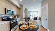 This thoughtfully designed single-floor residence showcases a contemporary aesthetic throughout, with light wood flooring, neutral tones, and recessed lighting creating a clean and welcoming ambiance. The open-concept layout seamlessly connects the kitchen, dining, and living areas, making the space feel expansive and functional. The kitchen is outfitted with stainless steel appliances, including a gas range, built-in microwave, and refrigerator, complemented by light wood cabinetry and a tiled backsplash. A round dining table sits adjacent to the kitchen, offering a casual dining experience. The living room is bright and airy, anchored by large floor-to-ceiling windows and a sliding glass door that opens to a private balcony, filling the space with natural light and outdoor views. The bedroom features plush carpeting, dual windows with outdoor views, and direct access to a walk-in closet with generous shelving and hanging space. Two bathrooms offer light wood vanities with white countertops and clean, modern finishes. A versatile secondary room with carpeted flooring serves well as a home office or den. Throughout, the design balances modern style with practical livability.