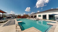 Located in Arlington, Texas, this outdoor amenity space centers around a generously sized resort-style swimming pool with crystal-clear blue water. The pool features a shallow entry area and is clearly marked for safety, offering a welcoming environment for residents of all ages. Surrounding the pool is an expansive, well-maintained deck finished in light-toned pavers, providing ample room for relaxation and socializing.

Numerous chaise lounges are arranged throughout the pool deck, both in open sun and beneath navy blue market umbrellas, offering flexible options for sunbathing or shaded lounging. A pergola structure at one end of the deck adds an architectural focal point and additional covered seating. Lush green artificial turf accents complement the hardscape, adding a touch of color and texture to the space.

Adjacent to the pool, a single-story clubhouse with white horizontal siding, large windows, and a dark shingled roof serves as the community's hub. The building opens directly onto the pool deck, creating a seamless indoor-outdoor connection. A privacy fence and black iron fencing enclose the perimeter, ensuring a secure and intimate setting. Under the broad Texas sky, this amenity area delivers a polished, community-focused outdoor retreat.