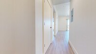 This modern single-floor unit showcases a clean, contemporary design throughout, with light wood-tone flooring and crisp white walls creating a bright, cohesive aesthetic from room to room. A well-appointed hallway with flush panel doors and recessed lighting guides you through the space, with an electrical panel neatly integrated into the corridor wall. The kitchen is outfitted with stainless steel appliances, including a range with a built-in overhead vent hood and a dishwasher, complemented by dark countertops and white cabinetry. The open layout connects the kitchen to the main living area, which is generously proportioned and filled with natural light. Multiple bedrooms offer large windows and sliding glass doors leading to private balconies, affording scenic outdoor views of surrounding trees and distant hills beneath open skies. Built-in closets with sliding doors provide ample storage in the bedroom spaces. The bathroom features a walk-in shower with white tile surround, a floating vanity with dark countertop and white cabinetry, black hardware accents, and a backlit mirror — delivering a polished, modern finish. Throughout the unit, the consistent use of light tones and natural light creates an airy, welcoming ambiance.