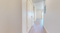 This modern single-floor unit showcases a clean, contemporary design throughout, with light wood-tone flooring and crisp white walls creating a bright, cohesive aesthetic from room to room. A well-appointed hallway with flush panel doors and recessed lighting guides you through the space, with an electrical panel neatly integrated into the corridor wall. The kitchen is outfitted with stainless steel appliances, including a range with a built-in overhead vent hood and a dishwasher, complemented by dark countertops and white cabinetry. The open layout connects the kitchen to the main living area, which is generously proportioned and filled with natural light. Multiple bedrooms offer large windows and sliding glass doors leading to private balconies, affording scenic outdoor views of surrounding trees and distant hills beneath open skies. Built-in closets with sliding doors provide ample storage in the bedroom spaces. The bathroom features a walk-in shower with white tile surround, a floating vanity with dark countertop and white cabinetry, black hardware accents, and a backlit mirror — delivering a polished, modern finish. Throughout the unit, the consistent use of light tones and natural light creates an airy, welcoming ambiance.