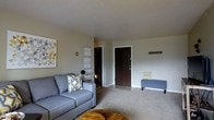 This single-floor space features an open living area finished in soft neutral tones, creating a calm and welcoming atmosphere. The living room is carpeted and comfortably proportioned, accommodating a sofa and coffee table with ease. Light gray walls serve as a versatile backdrop, complemented by decorative wall art that adds personality to the space. A dark wood-tone front entry door opens directly into the living area, which transitions to a tiled entryway, providing a clear delineation between the entrance and the carpeted living space. A wall-mounted coat hook near the entry offers practical storage. Adjacent to the main living area, a darkened hallway leads to additional rooms beyond. The overall design is straightforward and functional, with clean lines and minimal architectural detailing. The neutral color palette throughout allows for flexible interior styling, while the layout maximizes the usability of the available square footage. The space presents a practical and move-in ready environment suitable for everyday living.