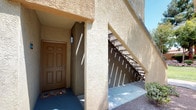 This single-story unit is set within a Southwest-inspired residential complex characterized by warm stucco exteriors, terra cotta accents, and mature landscaping featuring palm trees, desert shrubs, and manicured greenery. The grounds include decorative rock beds and open lawn areas, giving the community a relaxed, resort-like atmosphere.

The unit is accessed through a recessed, covered entryway framed by textured stucco walls, providing shade and a sense of privacy before entering. A staircase positioned adjacent to the entry indicates ground-floor placement within the multi-unit building.

Inside, the living area is bright and open, featuring light wood-style flooring and neutral wall tones that create a clean, versatile backdrop. A large sliding glass door dominates one wall, flooding the space with natural light and providing direct access to a private patio. The patio overlooks the beautifully maintained courtyard, offering views of tall trees, lush shrubs, and a community pergola structure.

The overall ambiance is light, airy, and comfortable, blending Southwest architectural character with a modern interior feel. The seamless indoor-outdoor connection through the patio enhances the living experience, making the surrounding landscape a natural extension of the interior space.