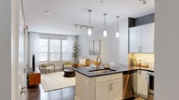 This thoughtfully designed one-bedroom, one-bathroom residence in Lansing, Michigan offers a modern open-concept layout that maximizes every square foot. The combined kitchen and living area serves as the heart of the home, where a sleek kitchen island with a dark granite countertop and undermount sink takes center stage. White shaker cabinetry, a tile backsplash, stainless steel appliances, and stylish pendant lighting complete the kitchen. The living area flows seamlessly from the kitchen, bathed in natural light from large windows dressed with blinds and sheer curtains, creating a bright and welcoming atmosphere. The bedroom is a comfortable retreat, featuring plush carpeting, generous natural light from dual windows with exterior views, and ample room for furnishings. The bathroom mirrors the kitchen's refined aesthetic with white cabinetry, dark granite countertops, a large vanity mirror, and a glass-enclosed shower. A dedicated in-unit laundry area with built-in shelving adds everyday convenience. Polished concrete flooring runs through the main living areas, adding an industrial-modern touch. A glass-paneled entry door connects to an exterior walkway, further enhancing the sense of openness throughout this well-finished residence.
