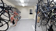 This indoor bicycle storage room is designed to provide a secure and organized space for cyclists. The room features a combination of wall-mounted bike racks along one side and a freestanding vertical rack system on the other, allowing for efficient use of space and accommodating a variety of bicycle styles and sizes. The floor is finished with a smooth, light-gray surface that is easy to maintain, while the neutral beige walls and drop ceiling with recessed fluorescent lighting create a clean and well-lit atmosphere. A windowed interior door provides access to the room, adding a measure of visibility and security. The space is thoughtfully laid out with a clear central aisle that allows for easy movement and retrieval of bicycles. With capacity for numerous bikes stored neatly in an upright or angled position, this room serves as a practical amenity for residents or building occupants who rely on cycling as their primary mode of transportation or leisure activity. The overall design prioritizes functionality and accessibility, making it a convenient and reliable storage solution.