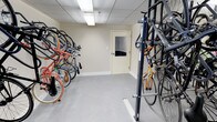 This indoor bicycle storage room is designed to provide a secure and organized space for cyclists. The room features a combination of wall-mounted bike racks along one side and a freestanding vertical rack system on the other, allowing for efficient use of space and accommodating a variety of bicycle styles and sizes. The floor is finished with a smooth, light-gray surface that is easy to maintain, while the neutral beige walls and drop ceiling with recessed fluorescent lighting create a clean and well-lit atmosphere. A windowed interior door provides access to the room, adding a measure of visibility and security. The space is thoughtfully laid out with a clear central aisle that allows for easy movement and retrieval of bicycles. With capacity for numerous bikes stored neatly in an upright or angled position, this room serves as a practical amenity for residents or building occupants who rely on cycling as their primary mode of transportation or leisure activity. The overall design prioritizes functionality and accessibility, making it a convenient and reliable storage solution.