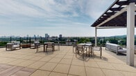 Located at 525 Lincoln St in Boston's vibrant Fenway neighborhood, this expansive rooftop terrace offers a compelling outdoor amenity space designed for relaxation and social gatherings. The terrace is finished with large-format, light-toned aggregate tiles that extend across the entire deck, lending a clean and polished aesthetic underfoot.

Multiple seating configurations are arranged throughout the space, including dining tables with modern metal chairs suited for al fresco meals and casual lounging areas with upholstered furniture. A pergola with a dark lattice roof provides a defined shaded retreat, while an additional covered overhang with a wood-paneled soffit adds architectural warmth to one end of the terrace.

A fully equipped outdoor kitchen station features multiple built-in gas grills set against a stone-clad counter, making the space ideal for communal cookouts. Decorative planters with lush greenery are positioned around the terrace, adding natural accents to the urban setting.

The rooftop is enclosed by a sleek black metal railing that frames sweeping, unobstructed views of the Boston skyline and surrounding tree canopy. Warm wood cladding on the rooftop access structure provides a contemporary contrast to the open sky above, completing a well-appointed outdoor destination.