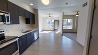 This contemporary single-floor space showcases a thoughtfully designed open-concept layout that seamlessly connects the kitchen and living areas. The kitchen is outfitted with dark navy cabinetry, light countertops, a stainless steel sink, and a dishwasher, offering a clean, modern aesthetic. The light-toned luxury vinyl plank flooring extends throughout the space, adding warmth and visual continuity.

The open living area is generously proportioned, benefiting from multiple large windows and a glass door that bathe the interior in natural light and provide views of the surrounding neighborhood. Ceiling fans are positioned in both the main living zone and an adjoining flex space, ensuring year-round comfort and airflow.

The overall ambiance is bright, airy, and minimalist, with neutral walls and recessed lighting complementing the natural light. The layout allows for flexible furniture arrangements, making the space adaptable to a variety of living configurations. The connection between the kitchen, dining, and living areas creates an inviting, functional environment well-suited for everyday living and entertaining alike.