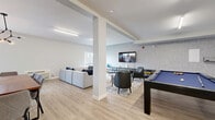 Located at 3641 Elliott Rd in West Kelowna, BC, this single-floor game room is a versatile and thoughtfully designed amenity space ideal for recreation and community gatherings. The open layout is anchored by light wood-look flooring, crisp white walls, and structural columns that define distinct activity zones without enclosing them.

At the heart of the space sits a full-size pool table with a blue felt surface, accompanied by tufted leather armchairs and framed landscape artwork on a textured accent wall. A wall-mounted flat-screen TV completes the entertainment zone, supported by a sleek media console and additional seating.

The lounge area features a generous sectional sofa arranged around round wooden coffee tables and a patterned area rug, with a window bringing in natural light. Adjacent to this is a dining zone with a large wooden table, upholstered chairs, and a modern branching pendant light fixture — suited for group meals or meetings.

A putting green adds a playful touch to the space, reflecting the room's commitment to varied recreational use. Bright linear ceiling fixtures ensure the entire area is well-lit throughout. The overall ambiance is modern and clean, balancing comfort with functionality in a spacious, multi-purpose setting.