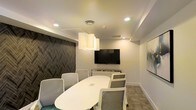 This single-floor conference space offers a refined and professional environment tailored for focused meetings and collaborative sessions. The room is anchored by a sleek, oval white conference table surrounded by a mix of cushioned white armchairs and mesh-backed task chairs, comfortably accommodating a sizable group. A large wall-mounted flat-screen display serves as the centerpiece for presentations and video conferencing, complemented by a credenza positioned beneath it for additional equipment or storage. A striking sculptural pendant light fixture hangs above the table, casting warm, even illumination that pairs with recessed ceiling lighting to create a well-lit, inviting atmosphere. The ceiling features subtle tray detailing, adding architectural depth to the space. One wall is adorned with a bold, textured chevron-patterned panel in dark tones, providing a sophisticated contrast to the surrounding neutral white walls. Patterned carpet tiles in muted tones anchor the space underfoot, while framed artwork on the adjacent wall adds a finishing decorative touch. The overall ambiance is clean, modern, and professional, making it well-suited for business meetings, presentations, or collaborative work sessions.