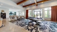 Located at 175 Main Street Market Place SE in Cartersville, Georgia, this single-floor community clubhouse offers a generously sized, open layout designed for both relaxation and social engagement. The space is anchored by soaring ceilings adorned with distinctive exposed wooden beams, creating a warm yet modern architectural character throughout.

The main living lounge area features large, comfortable sectional seating and accent armchairs arranged around a central coffee table, complemented by a wall-mounted flat-screen television flanked by bold, colorful abstract artwork. Adjacent to this, a dedicated game room houses a full-size billiards table set atop a striking geometric area rug, ideal for recreation and entertainment.

A dining zone with multiple tables and chairs provides a communal gathering space, while a sleek bar nook stands out with its vibrant teal hexagonal tile accent wall, open wooden shelving, pendant lighting, and a breakfast bar with leather seating — perfect for casual socializing.

Large glass doors and expansive windows throughout the space invite abundant natural light and offer views of the surrounding outdoor courtyard and community grounds. Light-toned tile flooring runs seamlessly across the space, enhancing the bright, airy ambiance. The overall design blends contemporary aesthetics with comfortable, inviting furnishings to create a welcoming environment for residents.