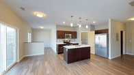 This single-floor space is designed with an open-concept layout that promotes a seamless flow between the kitchen and living areas. The kitchen serves as the centerpiece, showcasing rich dark wood cabinetry that provides ample storage alongside a suite of stainless steel appliances, including a French door refrigerator, range, and over-the-range microwave. A spacious central island with a built-in sink and light countertops offers both a functional workspace and a natural gathering point, illuminated by a trio of stylish pendant lights hanging overhead. A dedicated pantry area adds further storage convenience. Recessed lighting and flush-mount ceiling fixtures ensure the space is well-lit throughout. Light-toned hardwood flooring extends across the entire floor, lending warmth and continuity to the open layout. Neutral gray walls create a clean, understated backdrop that complements the contrast between the dark cabinetry and bright countertops. A raised half-wall at the edge of the kitchen subtly defines the transition into the adjacent living area, maintaining the open feel while providing a sense of spatial organization. The overall ambiance is modern, clean, and functional.