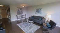 This well-appointed single-floor space showcases a seamless open-concept design that connects the living and dining areas in a cohesive, contemporary layout. The living area is anchored by a plush dark grey sofa accented with teal and charcoal throw pillows, complemented by a sleek glass-and-chrome coffee table set atop a luxurious shag area rug. A tufted accent chair adds an additional seating option, while a softly lit floor lamp contributes to the warm ambiance of the room. The dining area features a round glass table with elegant high-back grey chairs, creating an intimate setting ideal for casual meals. Dark hardwood flooring runs throughout the space, providing a rich contrast to the soft, neutral grey walls. Carefully selected wall art — including a dramatic cityscape print and a botanical close-up — adds personality and visual interest to the space. The interior design maintains a consistent modern aesthetic through the use of chrome accents, cool-toned textiles, and clean-lined furniture. The overall atmosphere is sophisticated yet inviting, with thoughtful attention to detail evident in every element of the décor.