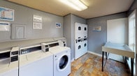 This single-floor communal laundry facility is designed to serve residents with practical, well-organized amenities. The space is finished with gray-painted walls, white trim, and multicolored stone-look tile flooring that adds a touch of warmth to the utilitarian setting. Overhead fluorescent lighting ensures the room is well-lit throughout.

The main laundry area is equipped with multiple front-loading and top-loading washing machines along one wall, complemented by stacked washer and dryer units along the adjacent wall, providing ample capacity for residents. A white utility folding table is positioned conveniently nearby, offering a dedicated surface for sorting and folding laundry.

A full-size Coca-Cola vending machine is situated near the entrance, providing refreshment options for residents during their visit. The room also features a utility sink, adding further functionality to the space.

Natural light filters in through several double-hung windows with white frames, offering views of the surrounding landscaped grounds. A standard white paneled entry door provides easy access. The overall layout is compact yet efficient, with all essential laundry amenities within easy reach, making this facility a convenient and practical resource for the residential community it serves.