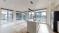 This contemporary urban residence is defined by its striking floor-to-ceiling windows, which wrap around the main living area and flood the space with natural light while offering expansive views of the surrounding cityscape. Light hardwood flooring runs consistently throughout, lending warmth and cohesion to every room.

The open-concept kitchen is a standout feature, anchored by a large island with a quartz countertop and an undermount sink with a matte black faucet. White shaker-style cabinetry, a classic subway tile backsplash, and a full suite of stainless steel appliances — including a refrigerator, range, and microwave — complete the kitchen's clean, modern aesthetic. A sleek linear pendant light hangs above the island, adding a sophisticated touch.

The living area benefits from corner window placement, creating a luminous, airy atmosphere with panoramic urban views on multiple sides. The bedrooms are generously proportioned, each featuring large windows with roller shades and ceiling fans, drawing in ample daylight and city views.

The bathrooms echo the home's contemporary design language, featuring white cabinetry with matte black hardware, quartz countertops, wood-look tile flooring, and a tiled tub-and-shower combination with coordinating black fixtures. Clean lines and neutral tones unify the space throughout.