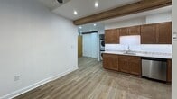 Located at 423 N 18th St in Richmond, VA, this efficiently designed single-floor unit blends modern finishes with distinctive industrial-inspired character. Exposed wooden ceiling beams run throughout the space, adding warmth and architectural interest to the bright, white-walled interior. The kitchen is a functional highlight, featuring rich wood cabinetry, granite countertops with an undermount sink, and a full suite of stainless steel appliances including a range, over-the-range microwave, and refrigerator. Recessed lighting illuminates the kitchen and living areas, complemented by a ceiling fan that adds comfort and style. The flooring transitions seamlessly between tile in the kitchen and wood-look planks throughout the remaining areas, creating a cohesive and contemporary aesthetic. A textured white masonry column serves as a striking architectural accent, adding a nod to the building's historic character. A dedicated in-unit laundry area with a stacked washer and dryer, tucked behind louvered doors, adds everyday convenience. The bedroom area features recessed lighting and a ceiling fan, with a built-in shelf providing additional utility. Clean white walls and crisp baseboards throughout contribute to a fresh, move-in-ready feel.